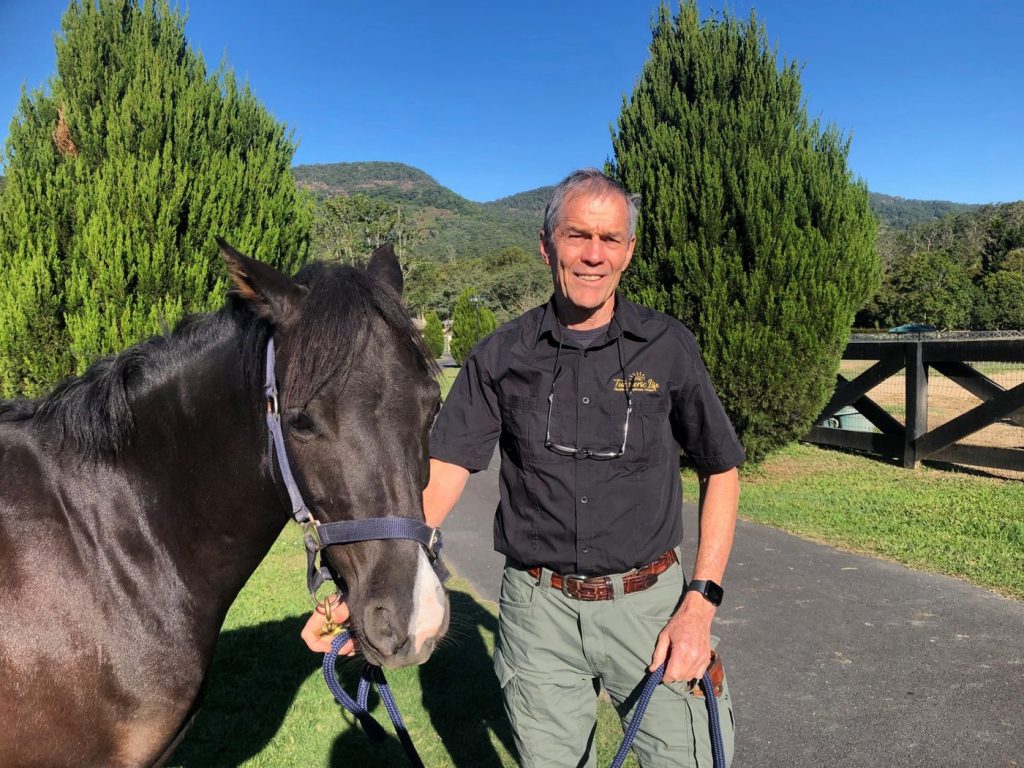 Dr Doug English, wearing a Turmeric Life shirt, standing with a black horse in front of green hills